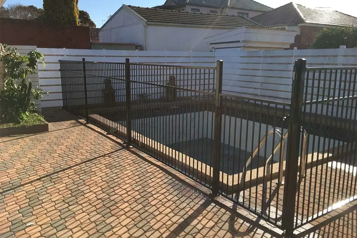 Black metal pedestrian gate within a pool enclosure fence beside paving