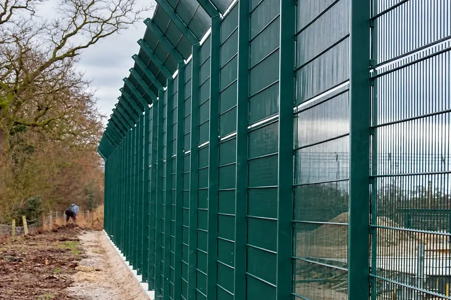 Tall green mesh perimeter fence with angled top along an energy facility boundary