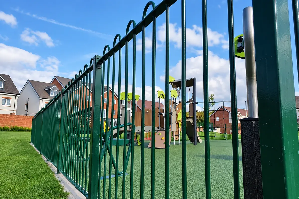 Green bow-top steel fence around a playground with play equipment beyond