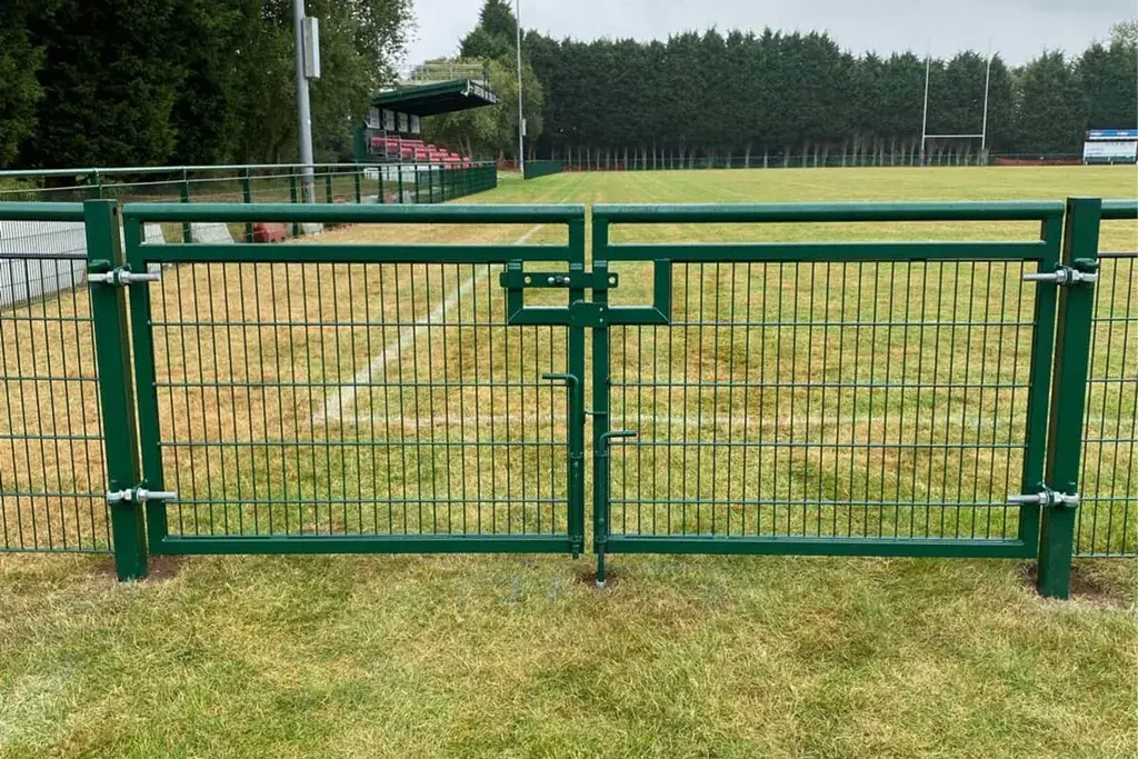 Green double-leaf mesh gate in a spectator rail beside a sports field