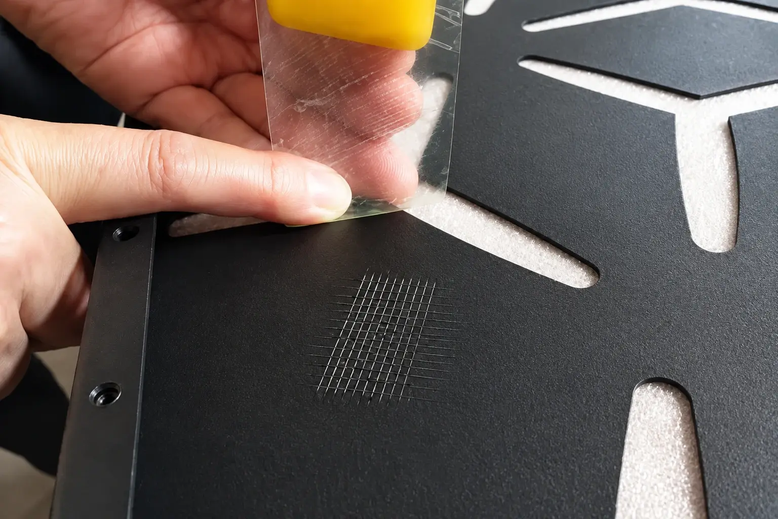 Hands applying tape to a cross-hatch cut on a dark coated metal sample