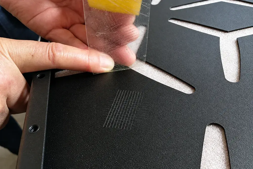 Hands checking a cross-hatch test area on a dark cut metal panel
