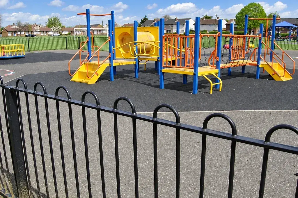 Black bow-top fence beside a playground with rubber play surfacing