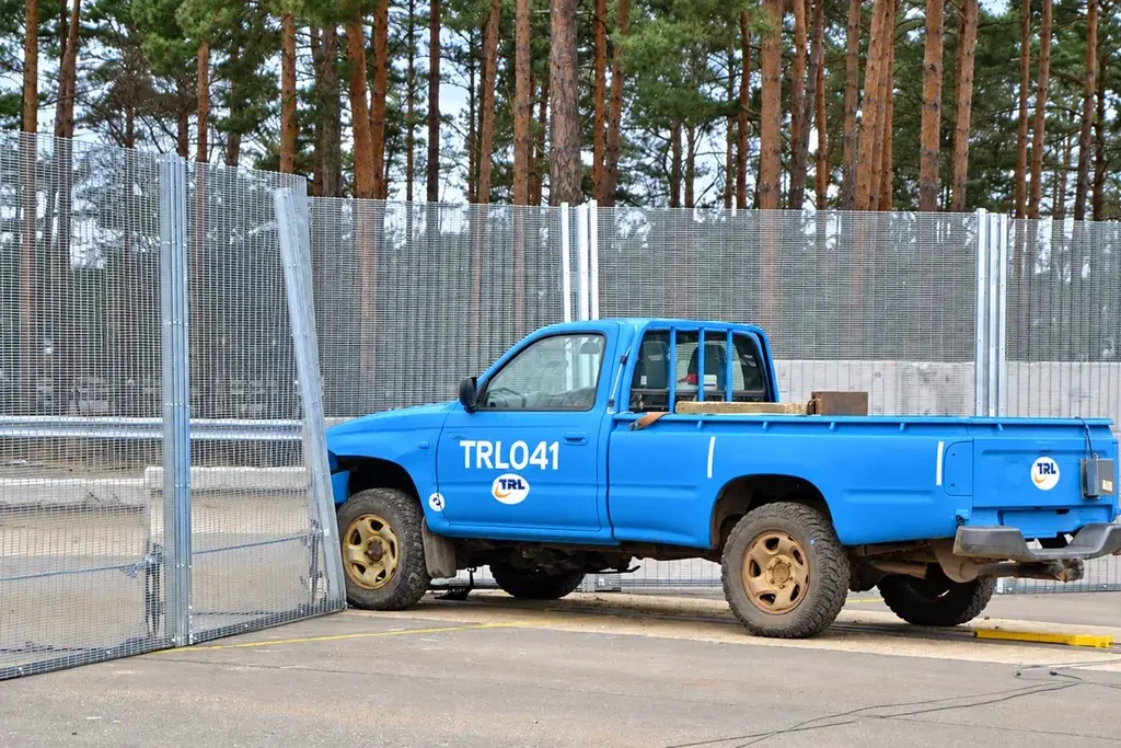 Blue pickup truck beside a tall security mesh fence at a test site