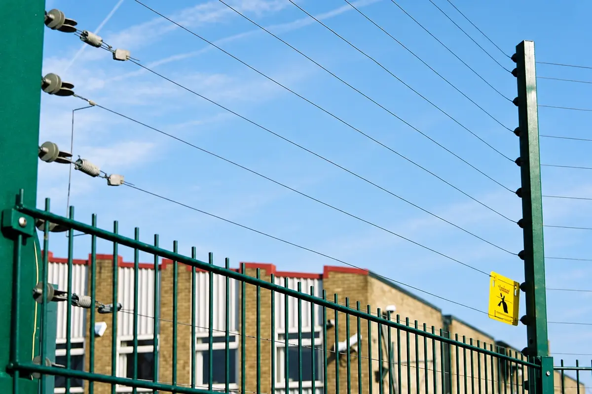 Electric fence wires and insulators above a metal fence with a warning sign