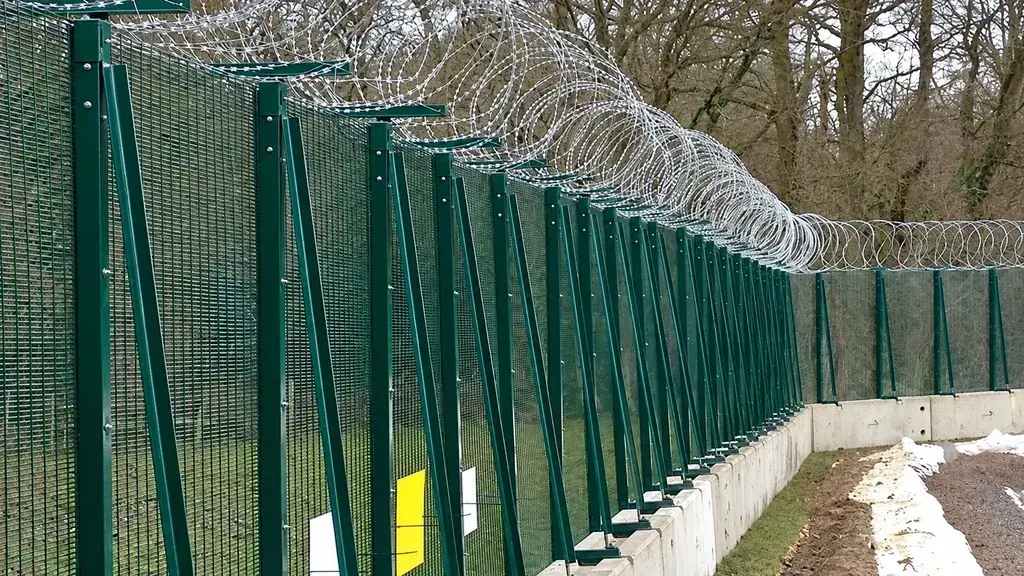 Green perimeter fencing with concertina wire on a concrete base