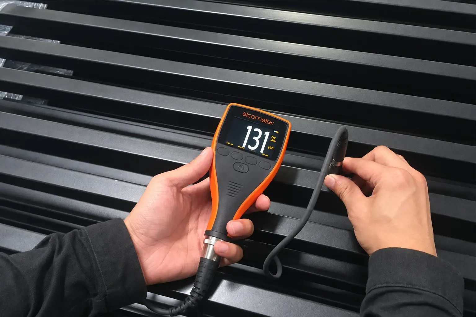 Handheld coating-thickness gauge and probe checking a black fence panel