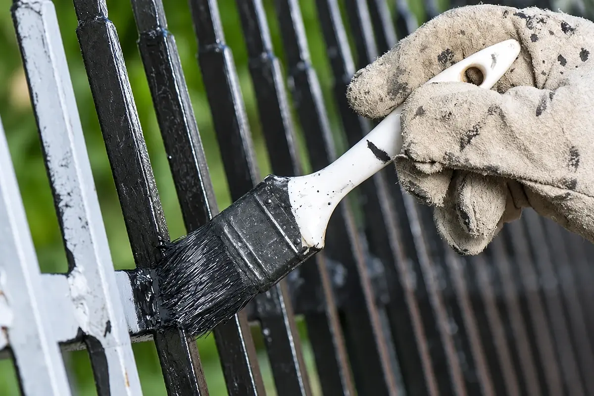 Gloved hand applying black finish to a steel fence bar with a brush