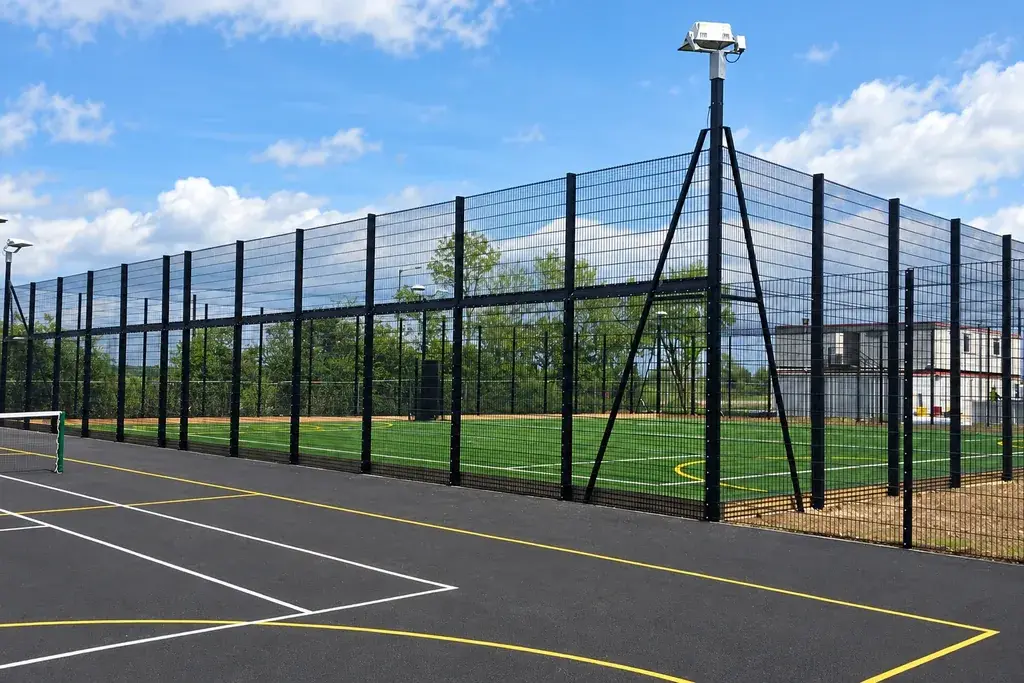 Tall sports court fencing beside a floodlight support and marked playing area